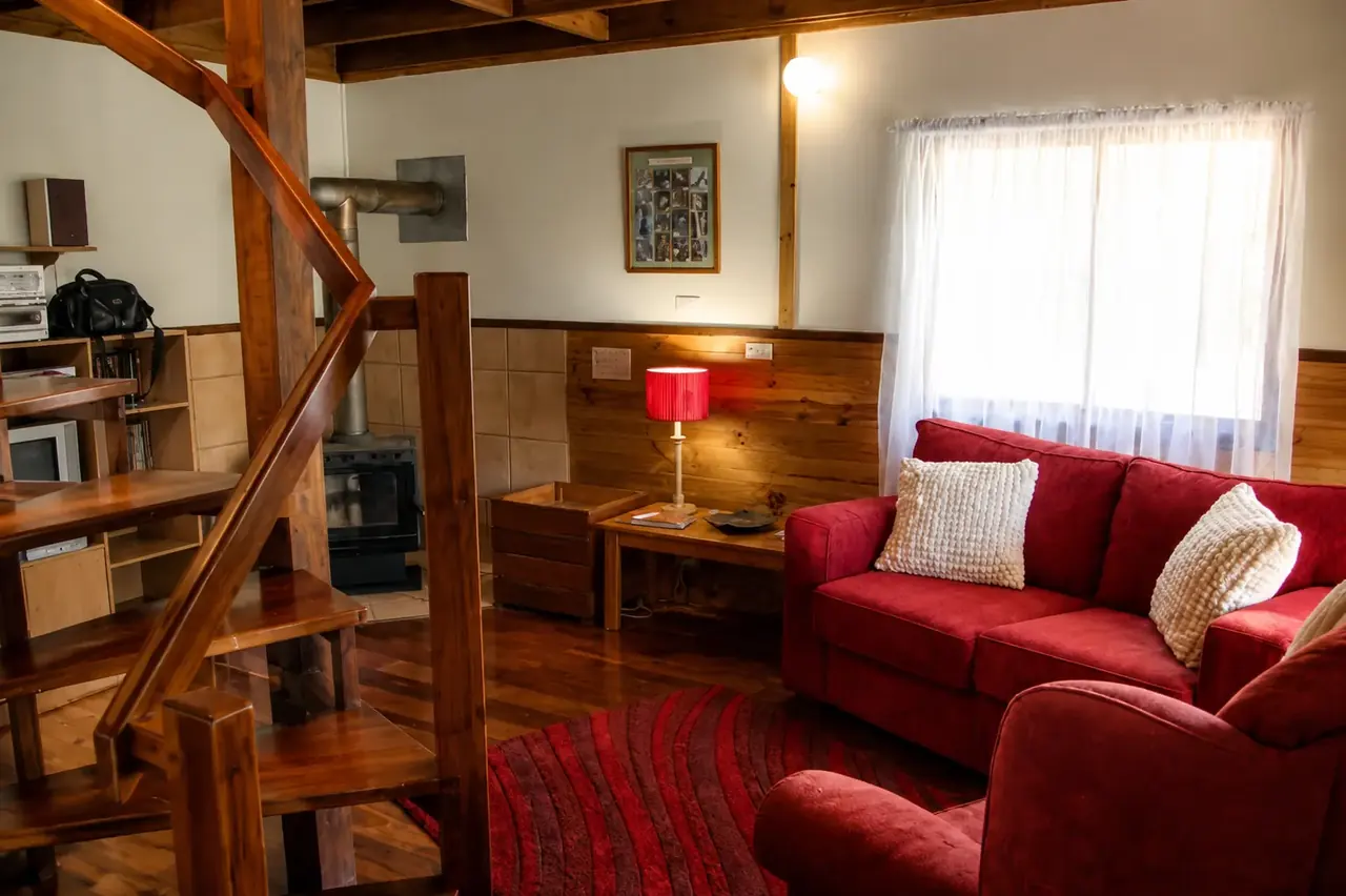 The lounge inside a chalet at Beyonderup Falls, a red sofa and armchair by the slow-combustion fire, the timber staircase alongside