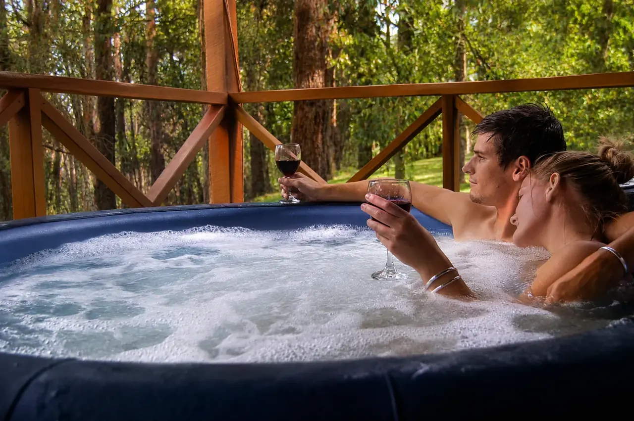 A couple relaxing with red wine in the private heated spa on a chalet verandah, forest beyond